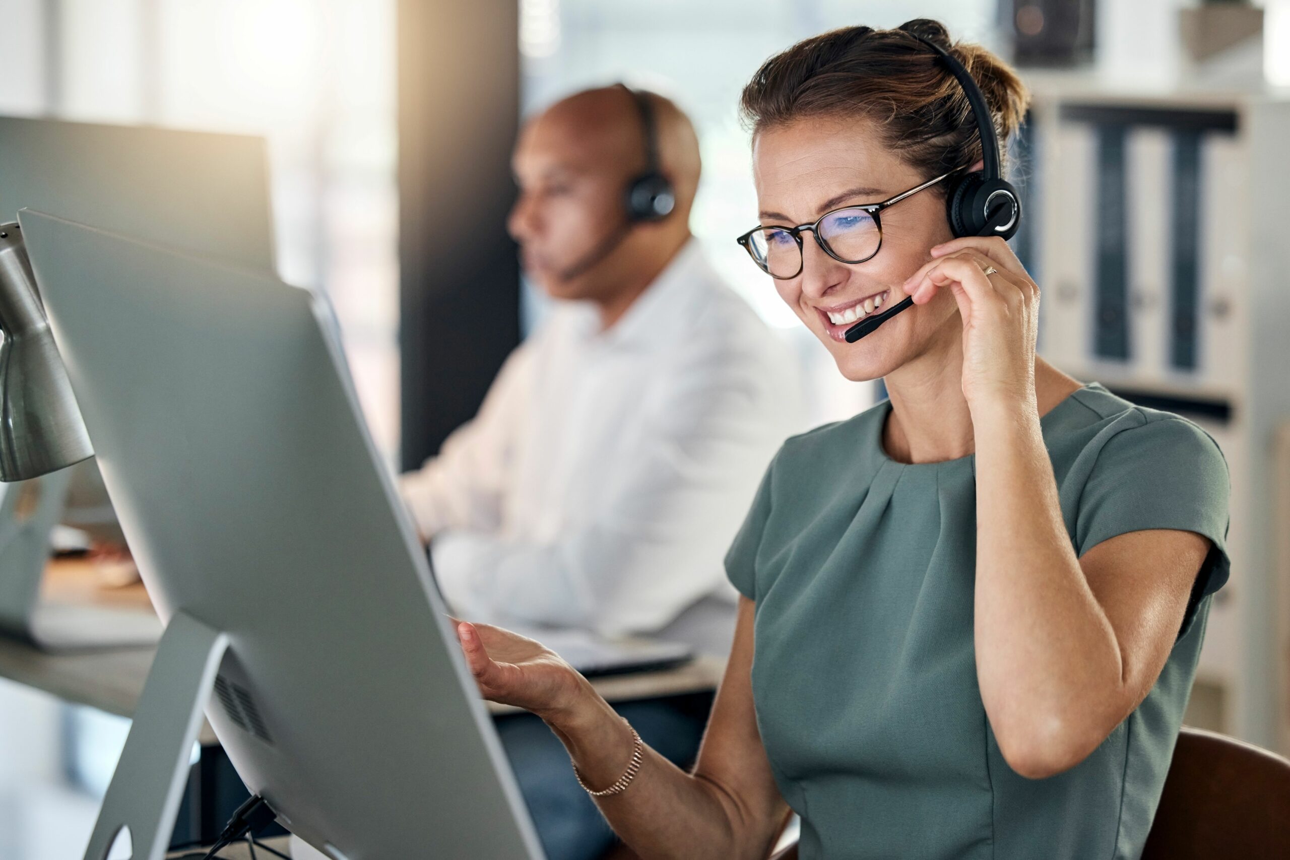 Eine Frau im Kundenservice telefoniert mit einer Person und schaut dabei auf ihren Computer. Im Hintergrund sitz ein weiterer Mitarbeiter.